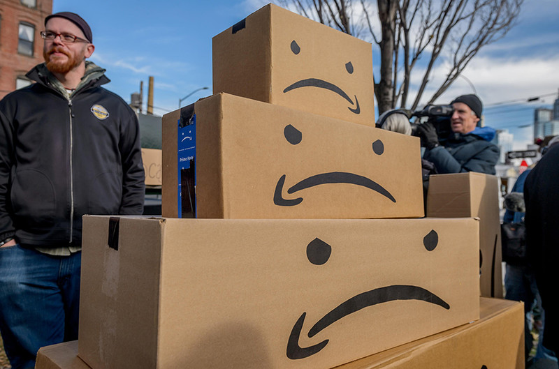 People protesting Amazon's plan to open headquarters in NYC in 2018. Amazon logo is made into sad faces on shipping boxes.